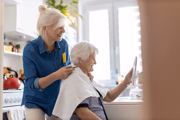 A smiling younger woman cuts the hair of an elderly woman, who is sitting in a kitchen and holding a mirror. Both appear happy and relaxed, with natural light coming through a window behind them.