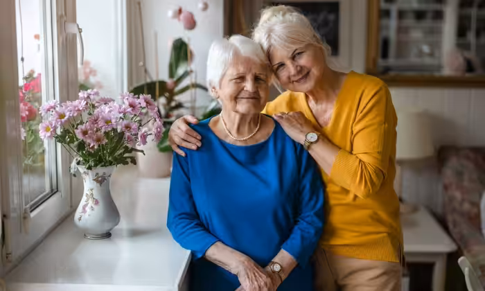 Two elderly women stand close together indoors. One, in a blue top, smiles gently while the other, in a yellow top, stands behind her, hugging her shoulders affectionately. A vase of pink flowers sits on the windowsill beside them.