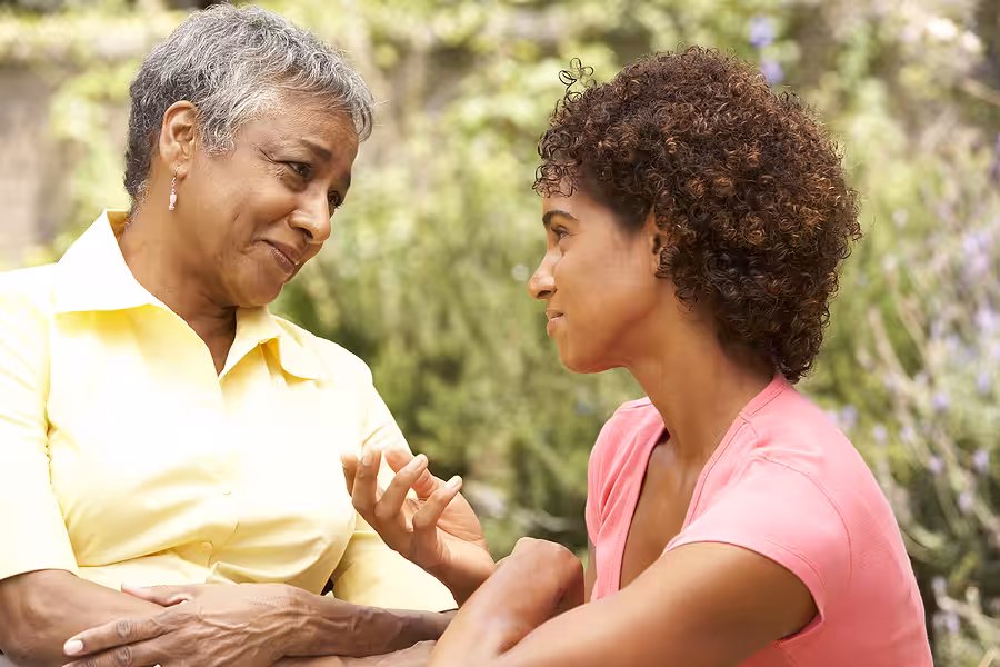 An older woman and a younger woman sit outdoors, facing each other and having a serious, heartfelt conversation. The older woman wears a yellow shirt, and the younger woman wears pink. Both appear engaged and empathetic.