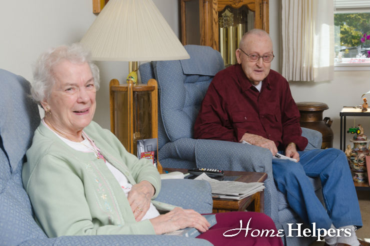 An elderly woman and man sit in armchairs in a cozy living room. The woman smiles at the camera, while the man looks on. A lamp, cabinet, and window are in the background. Home Helpers is written in the bottom right corner.