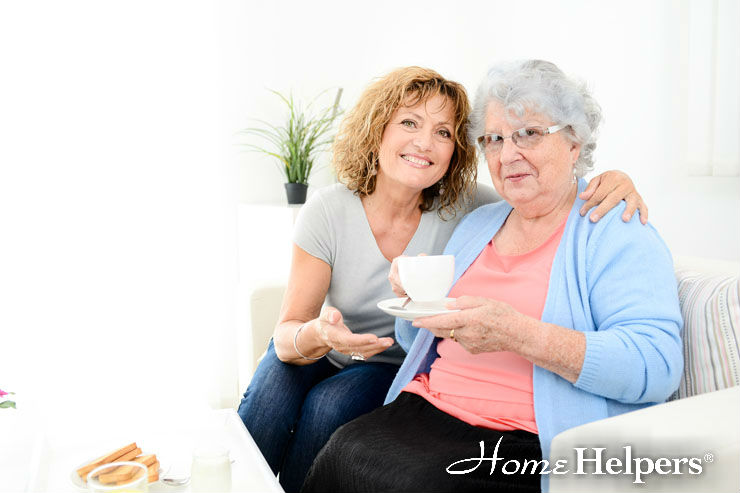 A smiling middle-aged woman sits beside and hugs an elderly woman holding a cup and saucer. They are seated on a couch, and the scene looks warm and friendly. The words Home Helpers appear in the bottom right corner.