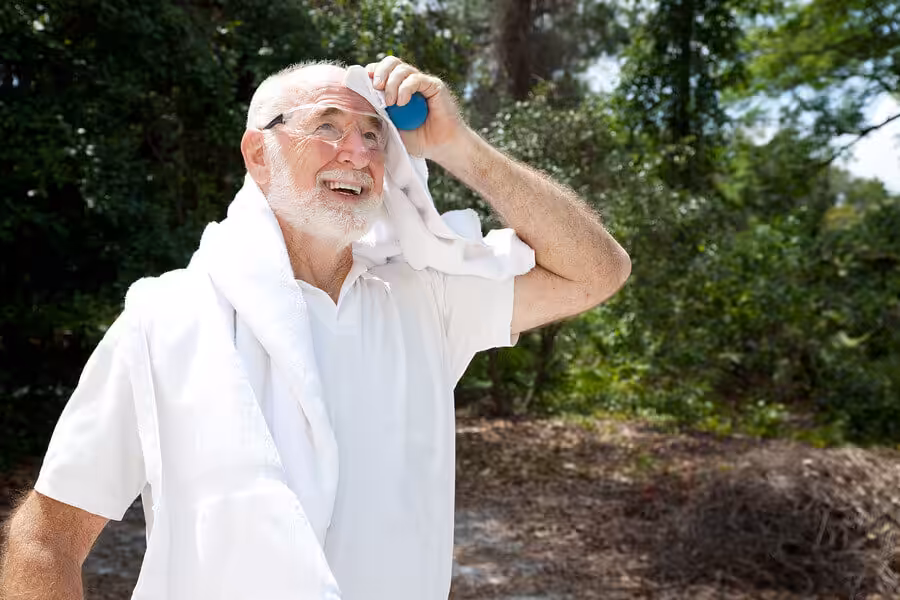 An elderly man outdoors wipes his forehead with a towel and holds a blue water bottle. He is smiling, wearing glasses and a white shirt, with trees and greenery in the background.