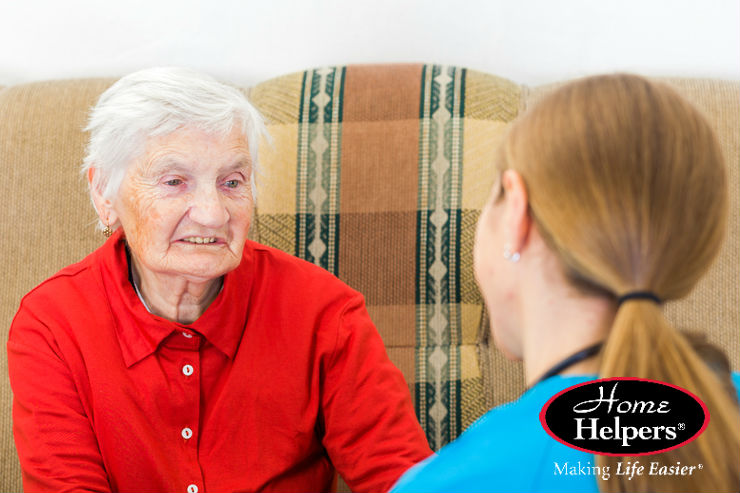 An elderly woman with white hair in a red shirt sits on a patterned sofa, smiling and holding hands with a caregiver. The Home Helpers logo and the phrase Making Life Easier appear in the bottom right corner.