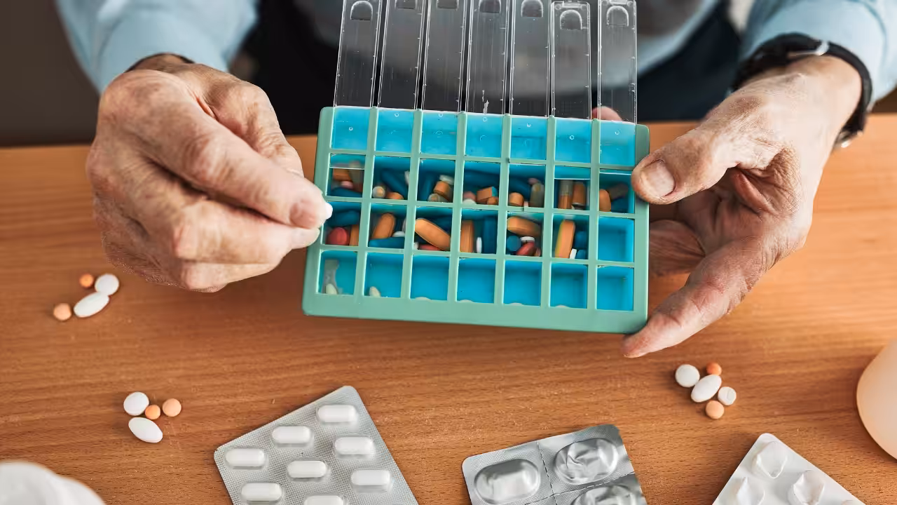 An elderly person’s hands holding a blue weekly pill organizer filled with various pills and capsules, with medication blister packs and loose pills on a wooden table.