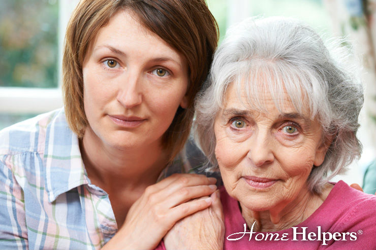 A younger woman gently holds the shoulder of an older woman with gray hair. Both look at the camera with calm expressions. The Home Helpers logo appears in the lower right corner.