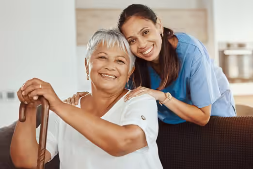 An older woman with short gray hair sits smiling with a cane, while a younger woman in scrubs stands behind her, resting her hands on the older womans shoulders. Both are smiling and looking at the camera.