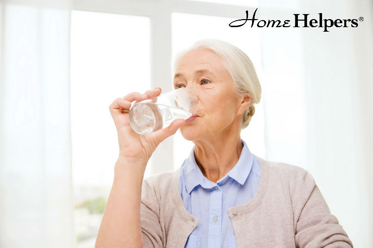 An older woman with gray hair, wearing a light blue blouse and beige sweater, drinks a glass of water. The background is bright, and the Home Helpers logo appears in the top right corner.