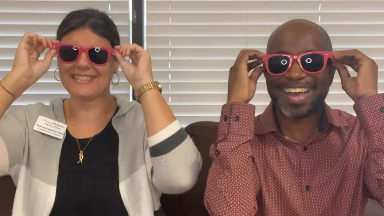 Two people sitting on a couch, smiling and putting on matching red sunglasses. The woman on the left has dark hair pulled back and wears a white sweater; the man on the right has a beard and wears a red patterned shirt.