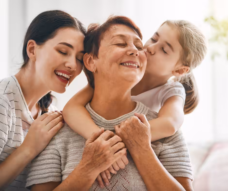 Three generations of women smile together; a young girl hugs and kisses an older woman on the cheek, while a younger woman stands beside them, smiling with eyes closed. All appear happy and affectionate.