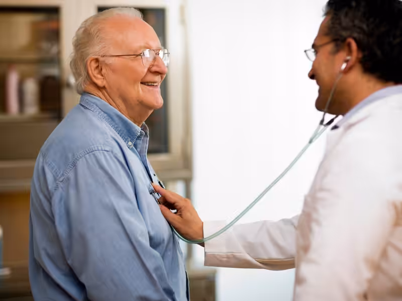 A doctor uses a stethoscope to listen to the chest of a smiling elderly man during a medical examination in a bright clinic room.