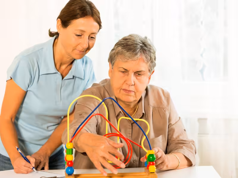 An older woman moves beads on a wire maze toy while a younger woman in a blue shirt observes and takes notes, possibly during a therapy or cognitive assessment session.