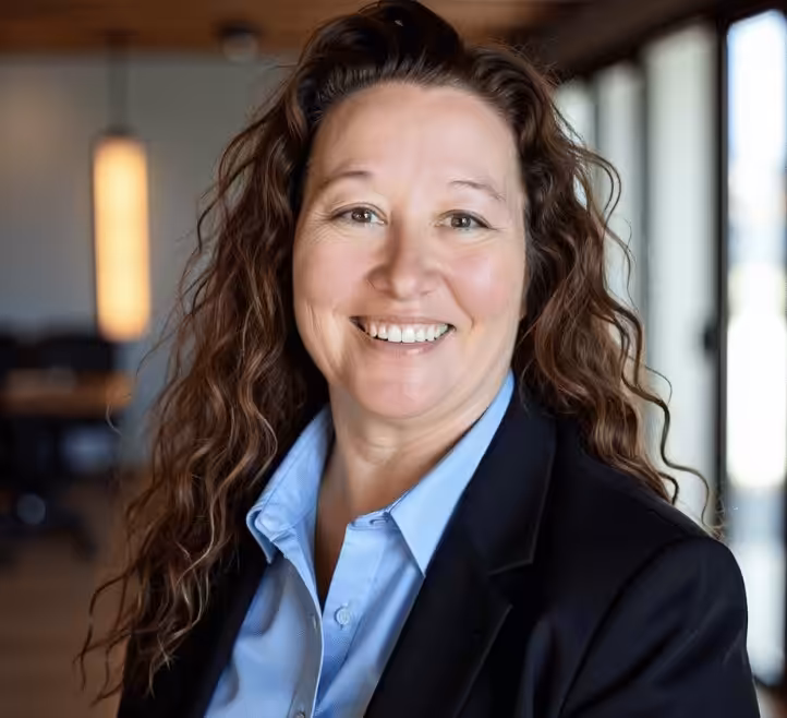 A woman with long, wavy brown hair, wearing a black blazer and light blue shirt, smiles at the camera in a modern office setting with large windows and warm lighting.