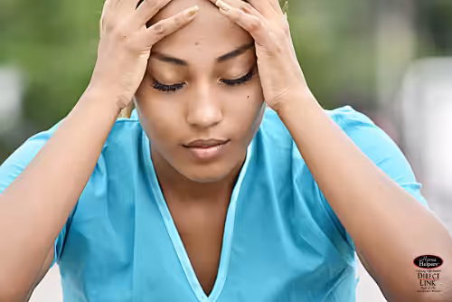 A young woman in a blue shirt sits with her eyes closed, holding her head in her hands, appearing stressed or overwhelmed. The background is blurred, emphasizing her facial expression and body language.
