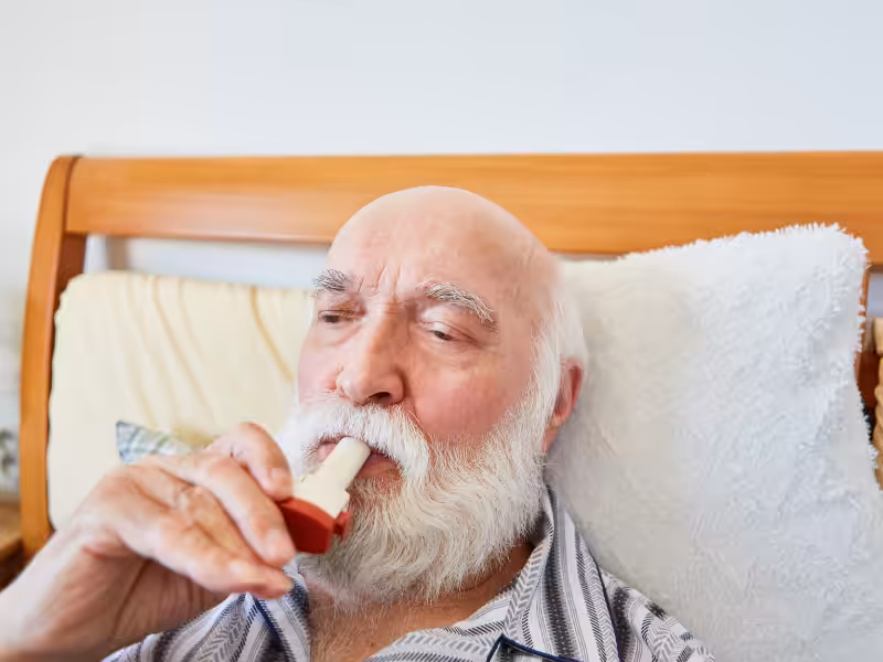 An elderly man with a white beard is sitting in bed, using an inhaler. He is wearing striped pajamas and is resting his head against a white pillow with a wooden headboard behind him.