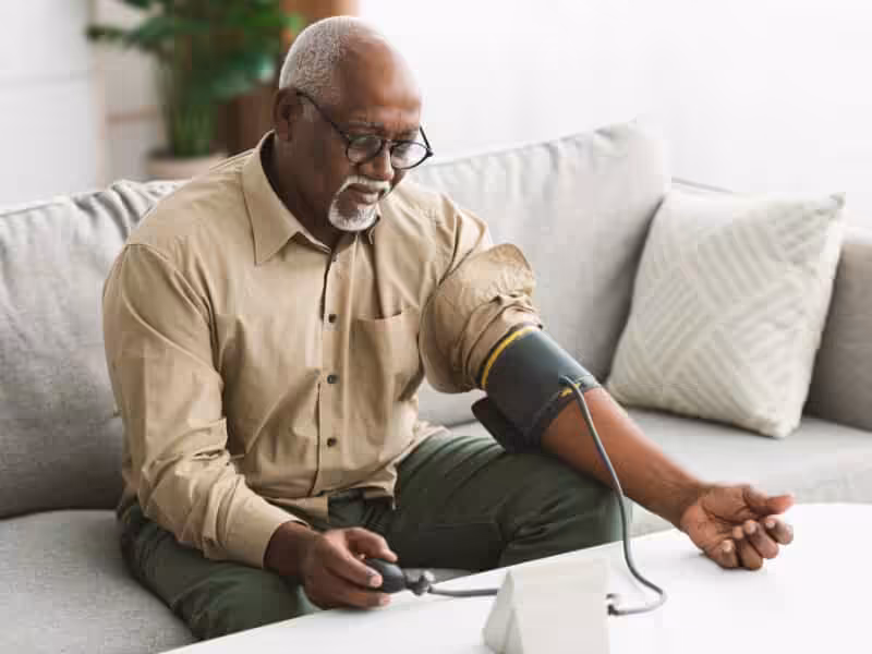 An older man sits on a couch and checks his blood pressure using a digital monitor, with the cuff wrapped around his upper arm and his shirt sleeve rolled up.