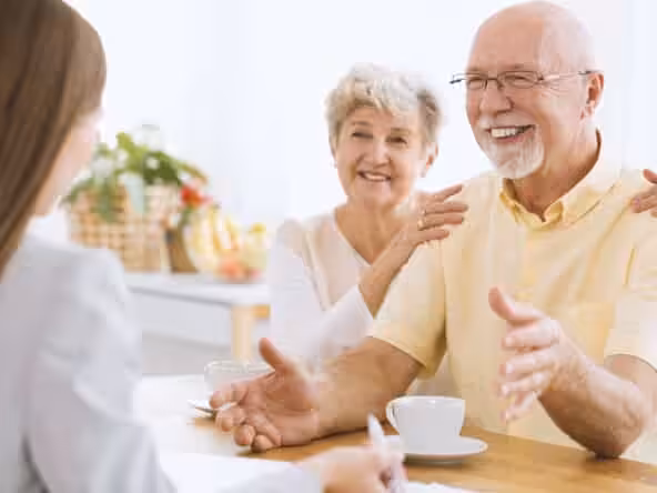 An elderly couple sits at a table with coffee cups, smiling and talking to a woman facing them. The setting is bright and cheerful, with fruit baskets in the background.