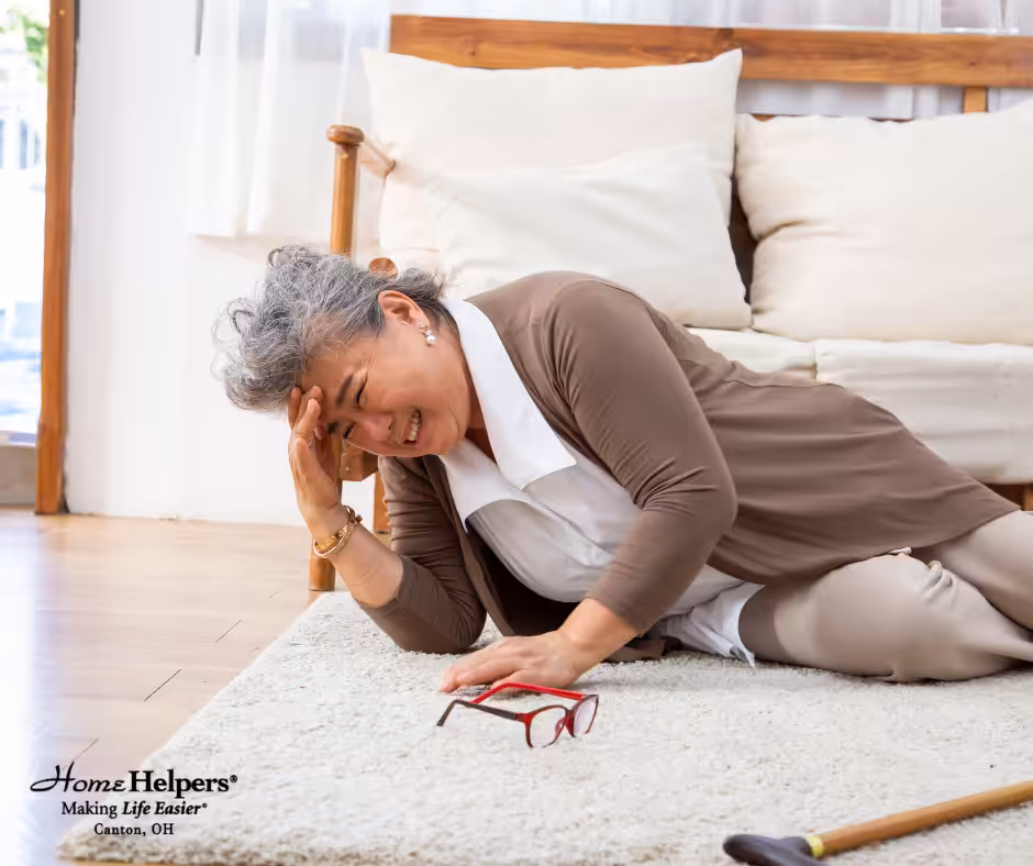 An older woman lies on a carpeted floor next to a walking cane and glasses, appearing to be in discomfort. She is holding her head and side, possibly after a fall, with a couch and window in the background.