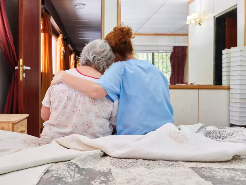 Two women sit side by side on a bed, one elderly and one younger, with their arms around each other, seen from behind in a warmly lit room.