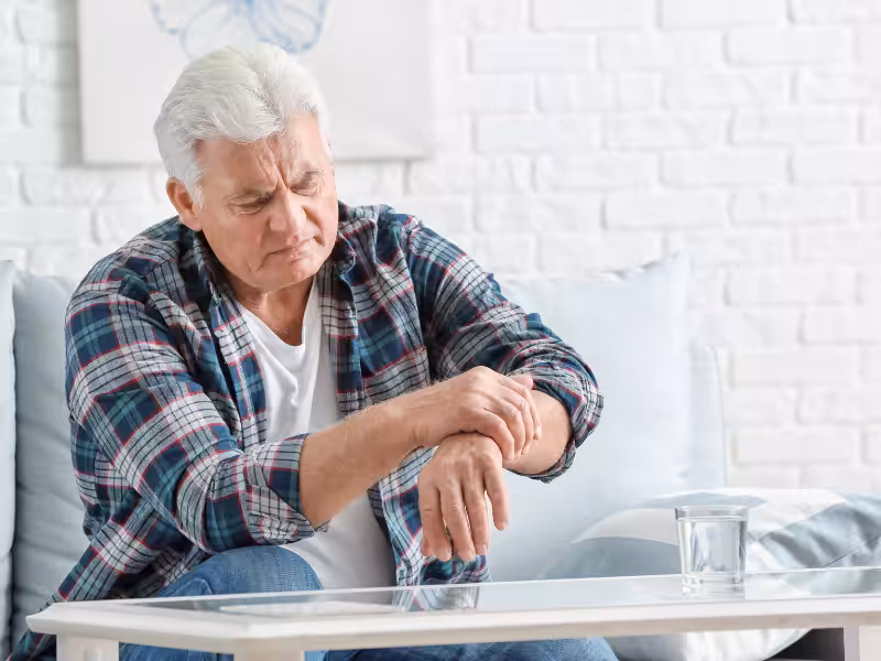 Older man sitting on a couch, looking at and touching his painful wrist with a concerned expression, a glass of water on the table in front of him.