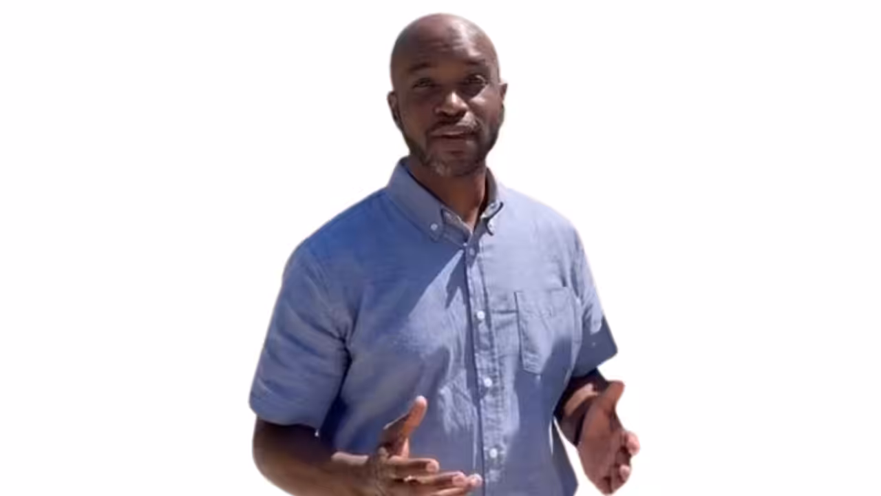 A man in a light blue button-up shirt stands against a white background, gesturing with his hands as if explaining or presenting something.