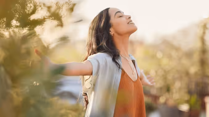 A woman stands outdoors with her arms outstretched, eyes closed, and a peaceful smile, enjoying the sunlight and fresh air among greenery.