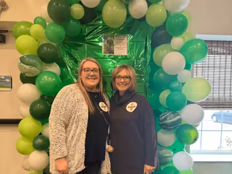 Two women smiling and standing in front of a green and white balloon arch indoors. Both wear glasses and dark tops with round badges. Sunlight streams through a window in the background.