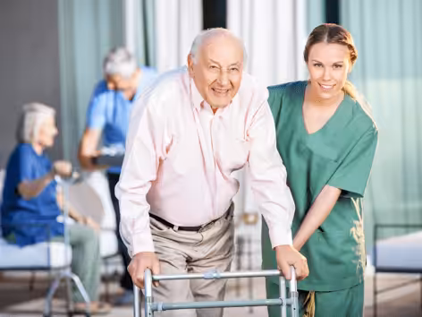 Elderly man using a walker is assisted by a smiling healthcare worker in green scrubs. In the background, two seniors and another caregiver interact in a bright, indoor setting.