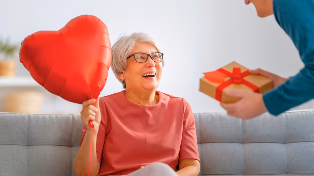 An older woman with gray hair and glasses smiles while holding a red heart-shaped balloon. A person is handing her a gift wrapped in brown paper with a red ribbon. They are sitting on a light-colored couch.