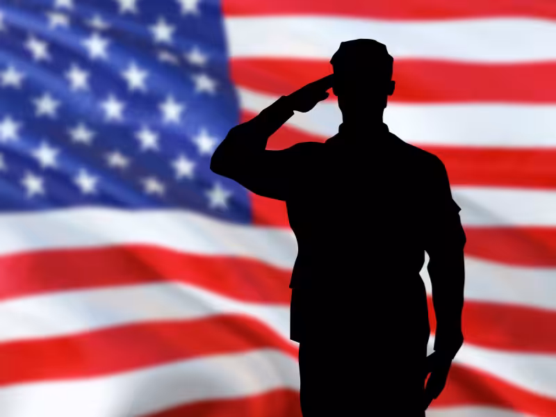 Silhouette of a person in military uniform saluting in front of a large, blurred American flag.