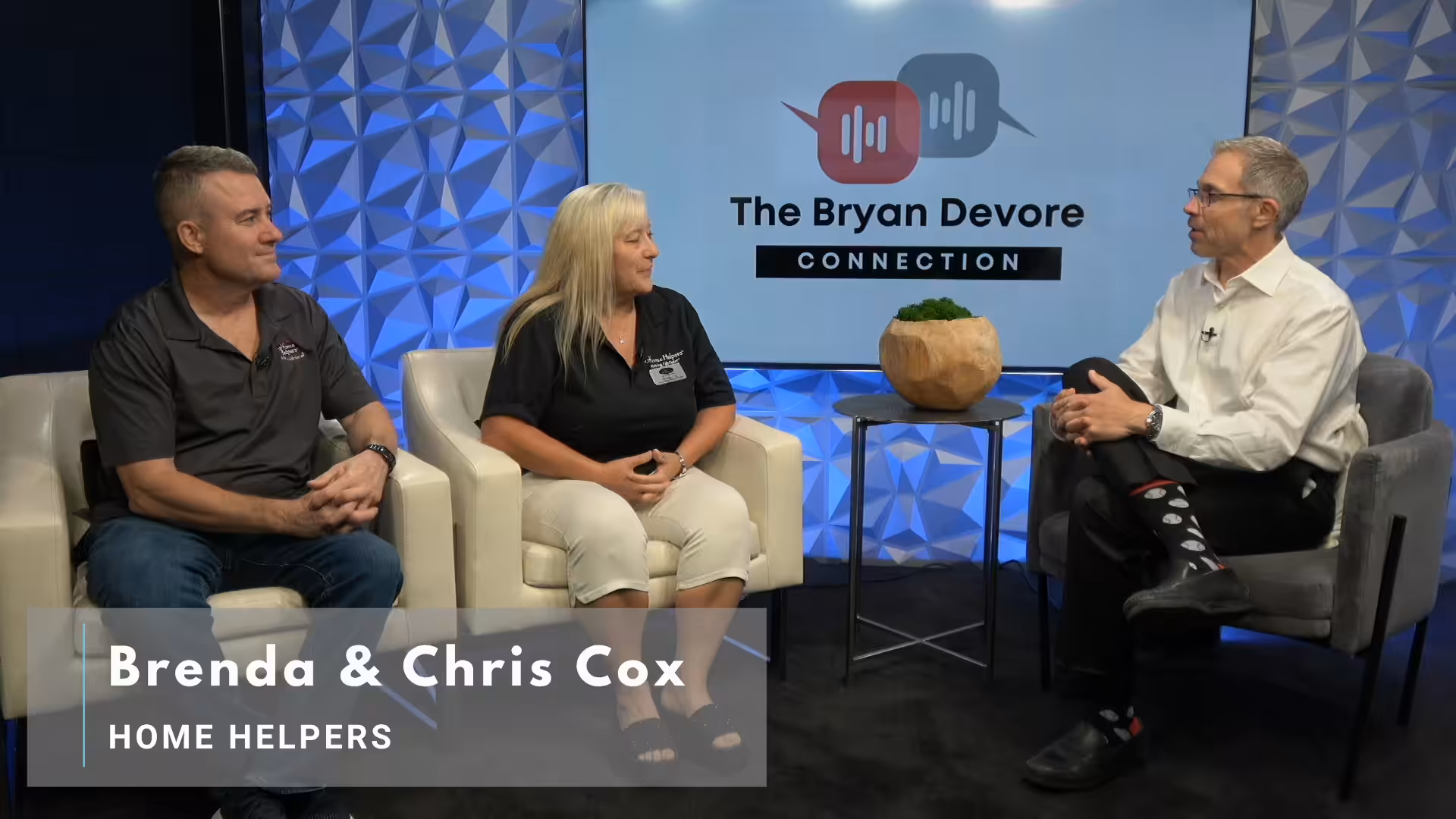 Three people are having a discussion in a studio. A man and woman, labeled as Brenda & Chris Cox from Home Helpers, sit on white chairs, while a host sits across from them. Behind them is a screen reading The Bryan Devore Connection.