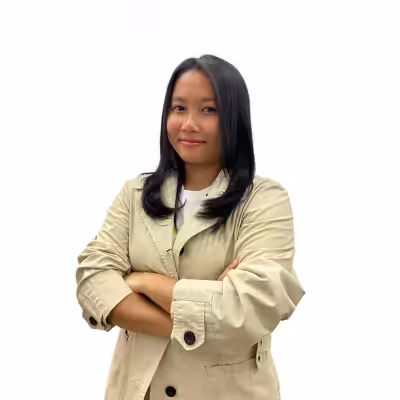 A person with long black hair, wearing a beige jacket and a white shirt, stands with arms crossed and smiles slightly against a plain white background.