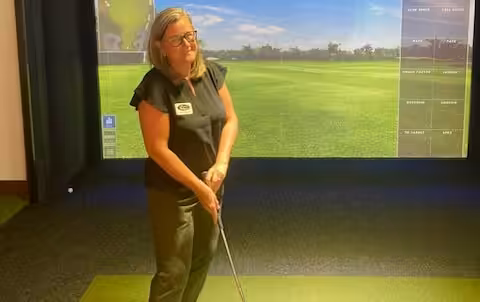 A woman with glasses stands indoors on a golf simulator mat, holding a golf club and preparing to swing, with a virtual golf course displayed on the screen behind her.