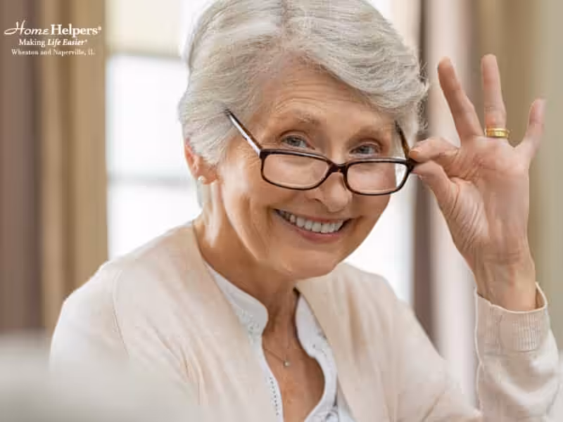 Smiling elderly woman with short gray hair and glasses, wearing a light cardigan, looks at the camera while slightly lifting her glasses with one hand. She appears cheerful and welcoming.