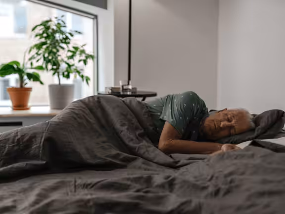 An older person wearing a green shirt with white dots is sleeping on their side in a bed with gray bedding. A window with potted plants is in the background.