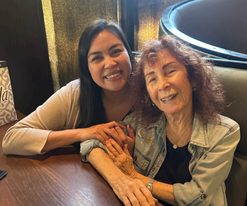 Two women sit closely together at a table, smiling warmly at the camera. The younger woman has long dark hair, while the older woman has curly hair and wears a light denim jacket. They hold hands affectionately.