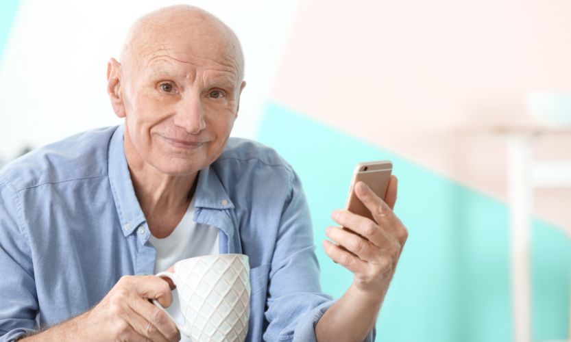 An older man with a bald head smiles while holding a white mug in one hand and a smartphone in the other, sitting in a bright room with pastel-colored walls.