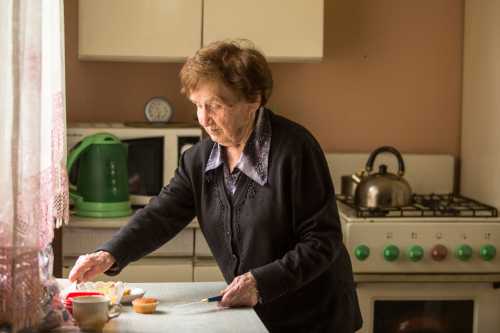 An elderly woman stands in a kitchen, cutting food at the counter. She is wearing a dark sweater and blouse. The kitchen has a stove, green kettle, microwave, and various small items on the counter. Sunlight comes through a window.