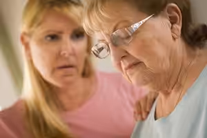 A younger woman gently comforts an older woman, who appears sad or thoughtful. The younger woman rests a hand on the older womans shoulder, showing concern and support.