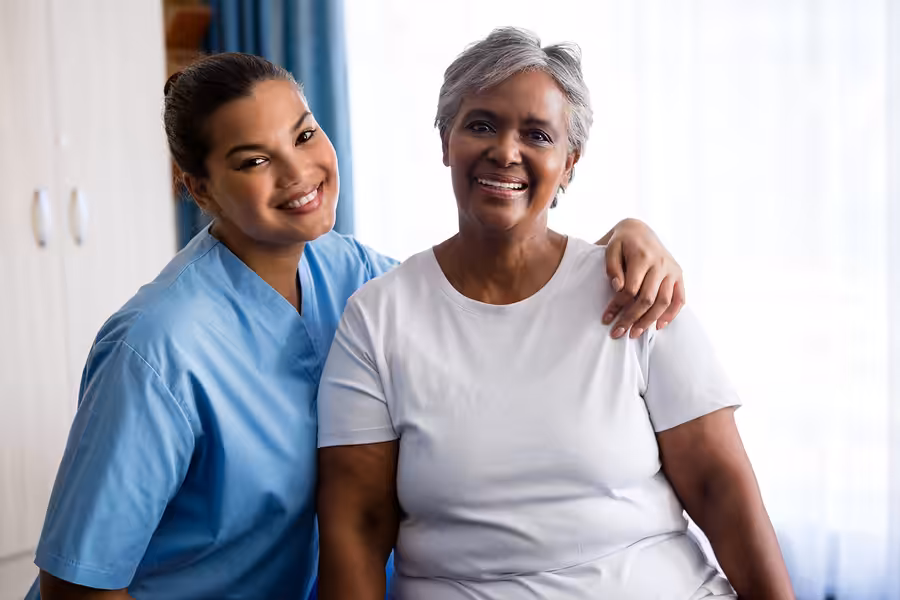 A smiling caregiver in light blue scrubs stands with her arm around an older woman in a white shirt. Both women are smiling and sitting indoors with natural light coming through a window.