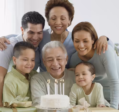 A smiling elderly man sits in front of a birthday cake with lit candles, surrounded by three adults and two children, all smiling and gathered closely around him in celebration.