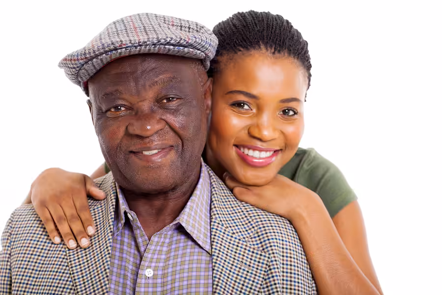 A smiling young woman embraces an older man from behind. The man wears a plaid flat cap and checked jacket, while the woman has braided hair and wears a green top. Both look happy against a white background.