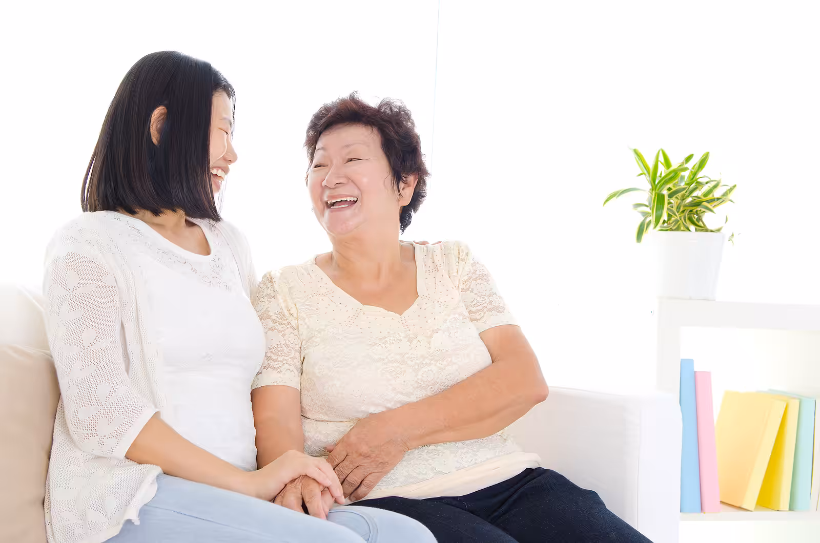Two women, one younger and one older, sit on a white couch holding hands and laughing together. A green potted plant and colorful books are on a shelf in the background. The mood is warm and joyful.