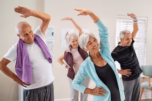 Four older adults in casual workout clothes are smiling and doing side stretches together in a bright room, participating in a group fitness class.