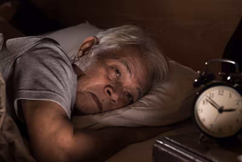 An elderly person with gray hair lies awake in bed at night, looking troubled. An old-fashioned alarm clock on a bedside table shows the time as 3:55. The room is dimly lit.