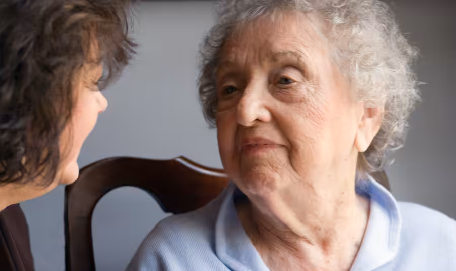 An elderly woman with short, curly gray hair and a light blue sweater looks at another woman sitting beside her, who is partially visible. They appear to be having a serious or thoughtful conversation indoors.