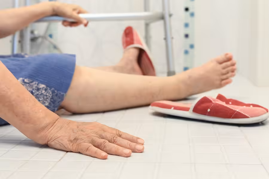 An elderly person wearing a blue dress and red slippers has fallen on the bathroom floor, with one hand on the ground and a walker nearby.