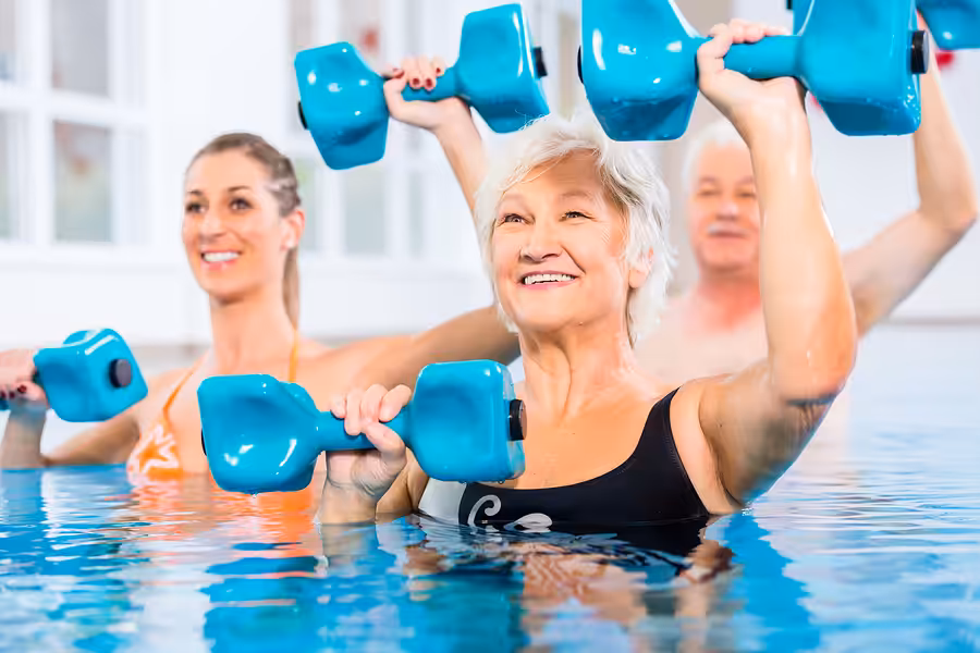 Three people in swimsuits smile while participating in a water aerobics class, holding blue foam dumbbells above their shoulders in a swimming pool. The group includes an older woman in front, with two others behind her.