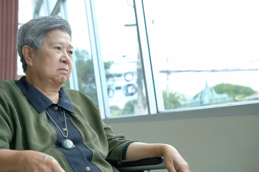 An elderly woman with short gray hair sits indoors near large windows, looking thoughtfully into the distance. She wears a green and navy top with a pendant necklace.