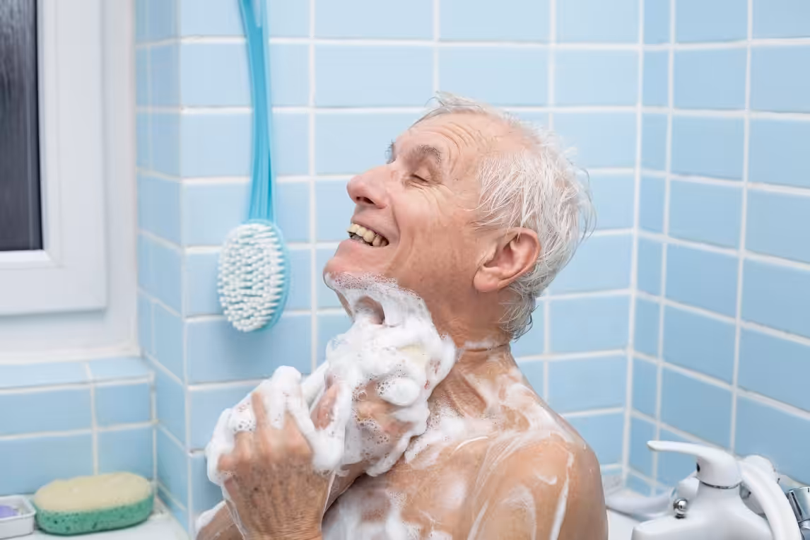 An elderly man with wet gray hair smiles while washing himself with soap in a tiled bathroom. He is covered in suds, and a blue bath brush hangs on the wall behind him.