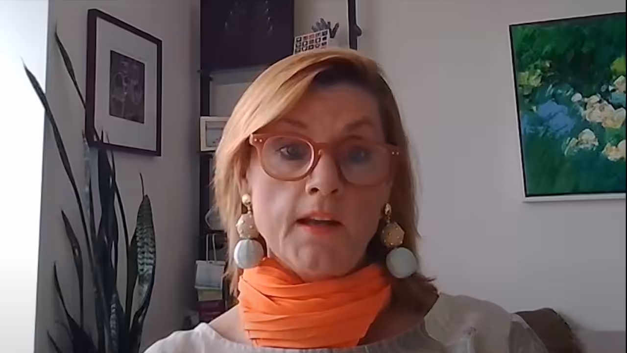 A woman with shoulder-length blond hair, wearing red glasses, large earrings, and an orange scarf sits indoors. Behind her are framed pictures, a green plant, and a painting on a white wall.
